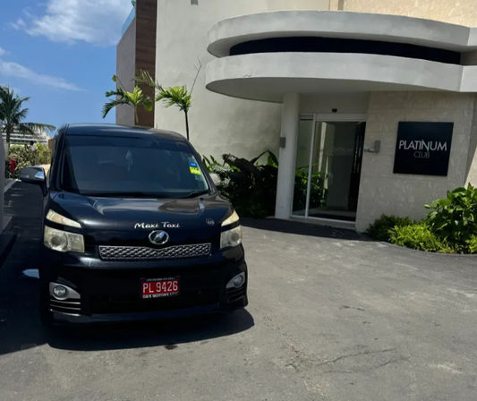 Black maxi-taxi shuttle van parked on a driveway outside a modern curved club entrance at a sunny tropical resort with palm trees and blue sky