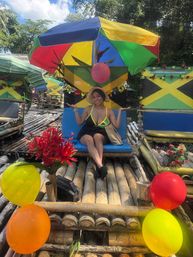 Smiling woman in a sunhat sits on a blue seat aboard a colorful bamboo raft decorated with Jamaican flag panels, balloons, red flowers and a multicolored umbrella on a sunny tropical river in Jamaica.
