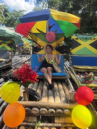 Smiling woman in a sunhat sits on a blue seat aboard a colorful bamboo raft decorated with Jamaican flag panels, balloons, red flowers and a multicolored umbrella on a sunny tropical river in Jamaica.