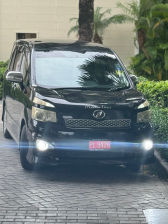 Front view of a black taxi minivan with headlights on parked on a palm-lined driveway outside a tropical hotel entrance
