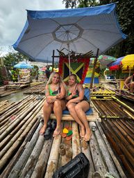 Two women smiling and flashing peace signs on a bamboo raft decorated with a Jamaican flag under a large umbrella during a tropical river rafting tour in Jamaica