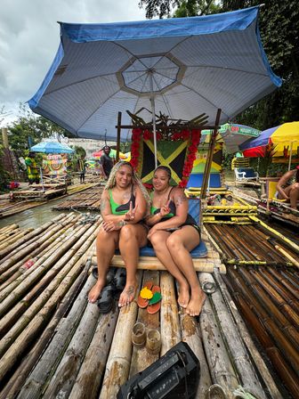 Two women smiling and flashing peace signs on a bamboo raft decorated with a Jamaican flag under a large umbrella during a tropical river rafting tour in Jamaica