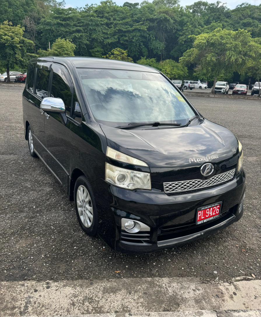 Sleek black minivan parked in a gravel parking lot, front three-quarter view with chrome mirrors and a tree-lined green background.