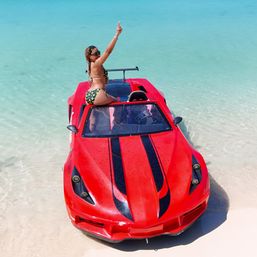 Woman in a green patterned bikini raising her arm while sitting on a red sports-car-style boat beached on clear turquoise water and white sand — tropical beach vacation.