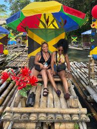 Two friends laughing on a bamboo raft under a colorful Jamaican-flag umbrella, surrounded by red tropical flowers and lush riverside greenery