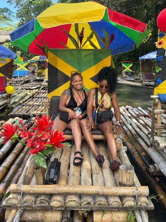 Two friends laughing on a bamboo raft under a colorful Jamaican-flag umbrella, surrounded by red tropical flowers and lush riverside greenery