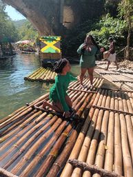 People enjoying bamboo raft ride on a clear tropical river beneath a stone arch, with a colorful raft painted with a Jamaican flag and bright umbrellas on the shore