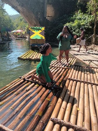 People enjoying bamboo raft ride on a clear tropical river beneath a stone arch, with a colorful raft painted with a Jamaican flag and bright umbrellas on the shore
