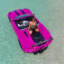 Aerial view of a woman in a green bikini sitting on a bright pink sports-car-style boat floating in clear turquoise shallow water.