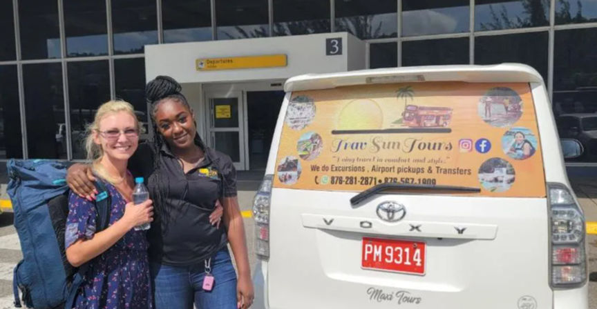 Two smiling travelers at an airport departures curb beside a white tour van with a colorful tropical decal, backpacks and bottled water ready for a sightseeing pickup.