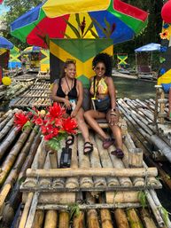 Two women relaxing on a bamboo river raft under a bright multicolored umbrella with a Jamaican flag backdrop, red tropical flowers and other rafts along a lush riverside — Jamaica bamboo rafting scene.