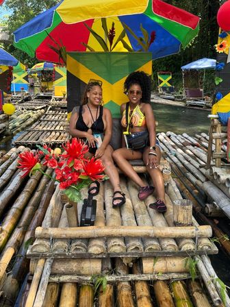 Two women relaxing on a bamboo river raft under a bright multicolored umbrella with a Jamaican flag backdrop, red tropical flowers and other rafts along a lush riverside — Jamaica bamboo rafting scene.