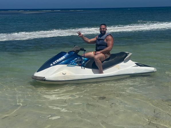 Smiling person in a life vest sitting on a blue-and-white jet ski in clear shallow tropical water, waving with gentle ocean waves and blue sky in the background.