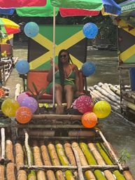 Person relaxing on a colorful bamboo raft with a Jamaican flag backdrop, umbrellas and birthday balloons on a tropical river.