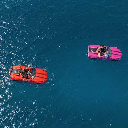 Aerial drone view of two bright convertibles (red and pink) floating on deep blue ocean water with people relaxing inside, a playful seaside scene.