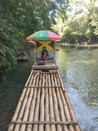 Person relaxing on a bamboo raft beneath a colorful umbrella and Jamaican flag, drifting along a clear tropical river framed by lush green trees