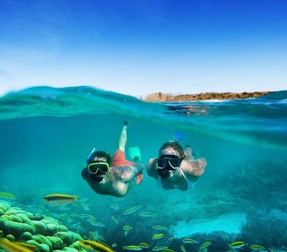 Over-under shot of two snorkelers exploring a vibrant tropical coral reef in crystal-clear turquoise water, surrounded by schools of small yellow fish.