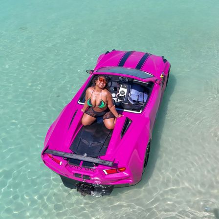 Smiling woman in a green bikini posing on a bright pink convertible sports car partially floating in crystal-clear turquoise shallow water, tropical beach vibe.