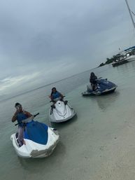 Three riders on jet skis in shallow clear tropical water by a sandy beach, one waving, with a moored sailboat nearby under an overcast sky