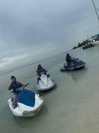 Three riders on jet skis in shallow clear tropical water by a sandy beach, one waving, with a moored sailboat nearby under an overcast sky