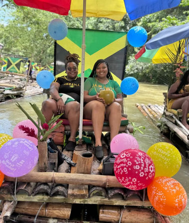 Two friends relaxing on a bamboo raft drifting down a Jamaican river under a colorful umbrella with a Jamaican flag backdrop, birthday balloons, tropical flowers, and a fresh coconut drink.