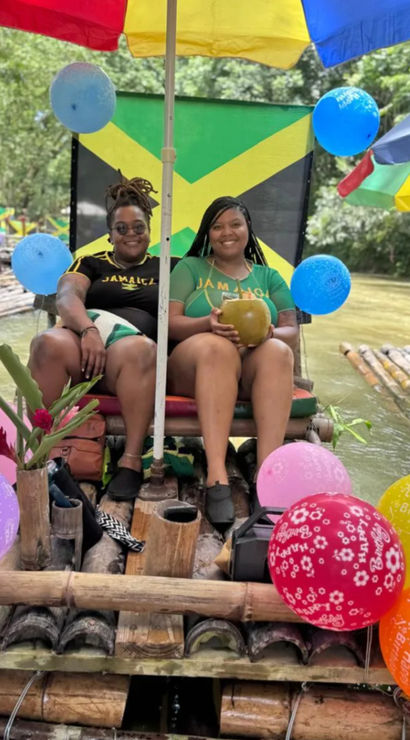 Two friends relaxing on a bamboo raft drifting down a Jamaican river under a colorful umbrella with a Jamaican flag backdrop, birthday balloons, tropical flowers, and a fresh coconut drink.