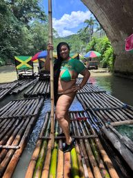 Smiling woman posing on a bamboo river raft in Jamaica, holding a pole and wearing green-and-yellow swimwear, with Jamaican-flag seats, tropical foliage, stone bridge and blue sky in the background.