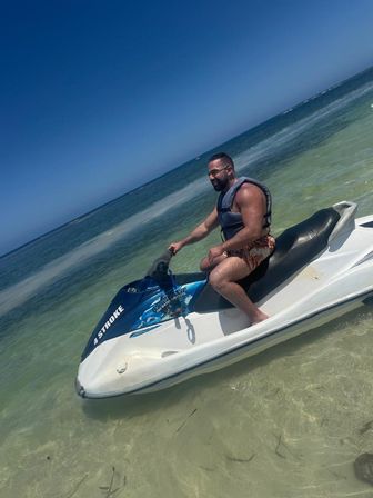 Person in a life vest seated on a white-and-blue jet ski in clear shallow turquoise water near shore under a sunny blue sky.