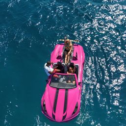 Aerial view of a bright pink convertible-style speedboat with three people enjoying a sunny day on sparkling deep-blue ocean water, one person lounging at the stern and waving.
