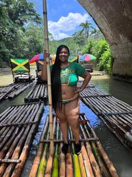 Smiling woman in Jamaica-themed swimwear stands on a bamboo raft holding a pole beneath a stone bridge on a lush tropical river, with colorful umbrellas and Jamaican-flag chairs in the background.
