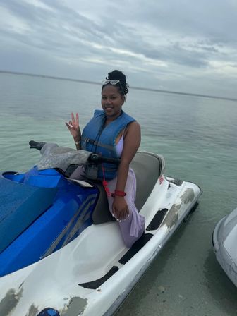 Smiling woman in a blue life jacket sitting on a blue-and-white jet ski in shallow clear ocean water, flashing a peace sign under a cloudy sky