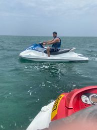 Person wearing a blue life vest riding a white-and-blue jet ski on green ocean water under a cloudy sky, with a red jet ski dashboard visible in the foreground — recreational water sports scene.