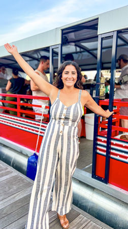 Smiling woman in gray-and-white vertical striped jumpsuit and sandals, arms raised on a dock beside a red pontoon boat at a marina