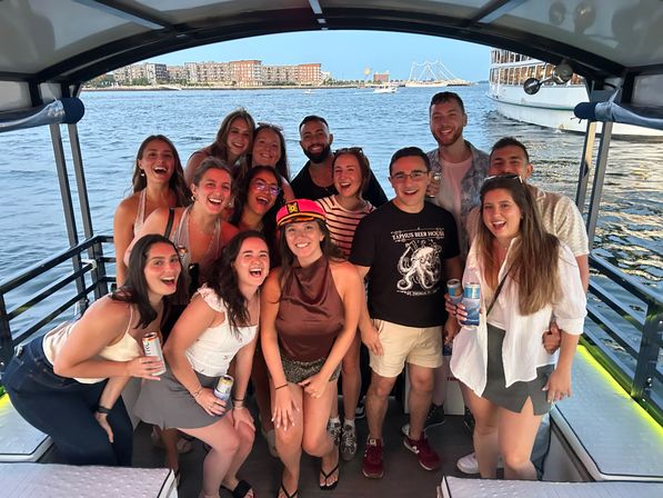 Group of friends on a covered party boat at sunset, laughing and holding drinks with a city waterfront skyline and harbor in the background.