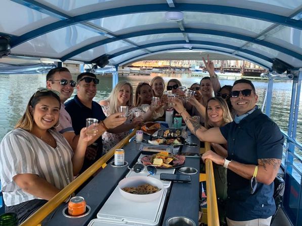 Cheerful group toasting with drinks on a covered pontoon party boat at a waterfront marina, central table with snacks and cans