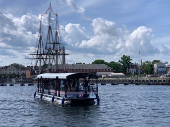 Covered harbor tour boat with passengers cruising past a moored three‑masted tall ship along a historic waterfront under a bright, partly cloudy sky.