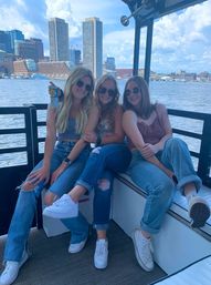 Three friends in sunglasses and jeans smiling on a boat bench with a canned drink, enjoying a sunny summer harbor cruise with an urban skyline and waterfront in the background.