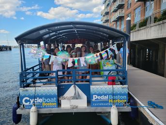 Group of people celebrating a birthday on a covered pedal boat at Boston Harbor dock, holographic "Happy Birthday" banner, gift bags and waterfront buildings in the background.