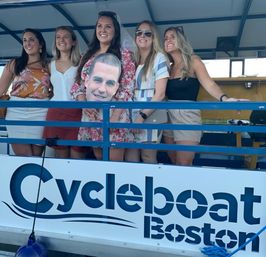 Five smiling women on a blue-deck Boston party pedal-boat, one wearing a veil and floral dress and holding a giant cardboard face cutout.