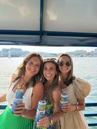 Three smiling friends on a boat holding canned drinks in blue koozies, posing against a sunny harbor marina with sailboats and waterfront buildings.