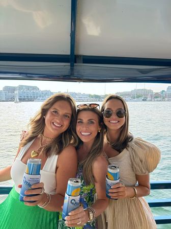 Three smiling friends on a boat holding canned drinks in blue koozies, posing against a sunny harbor marina with sailboats and waterfront buildings.