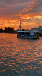 Fiery orange sunset over a coastal harbor, rippling water reflecting the sky, a pedal-powered tour boat near waterfront buildings with a tall mast silhouette.