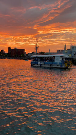Fiery orange sunset over a coastal harbor, rippling water reflecting the sky, a pedal-powered tour boat near waterfront buildings with a tall mast silhouette.