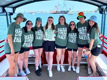 Smiling bachelorette group on a sunny boat cruise — eight friends in matching 'Here Comes the Party' tees with the bride in a 'Here Comes the Bride' shirt, posing on a harbor with a large tall ship and blue water in the background.