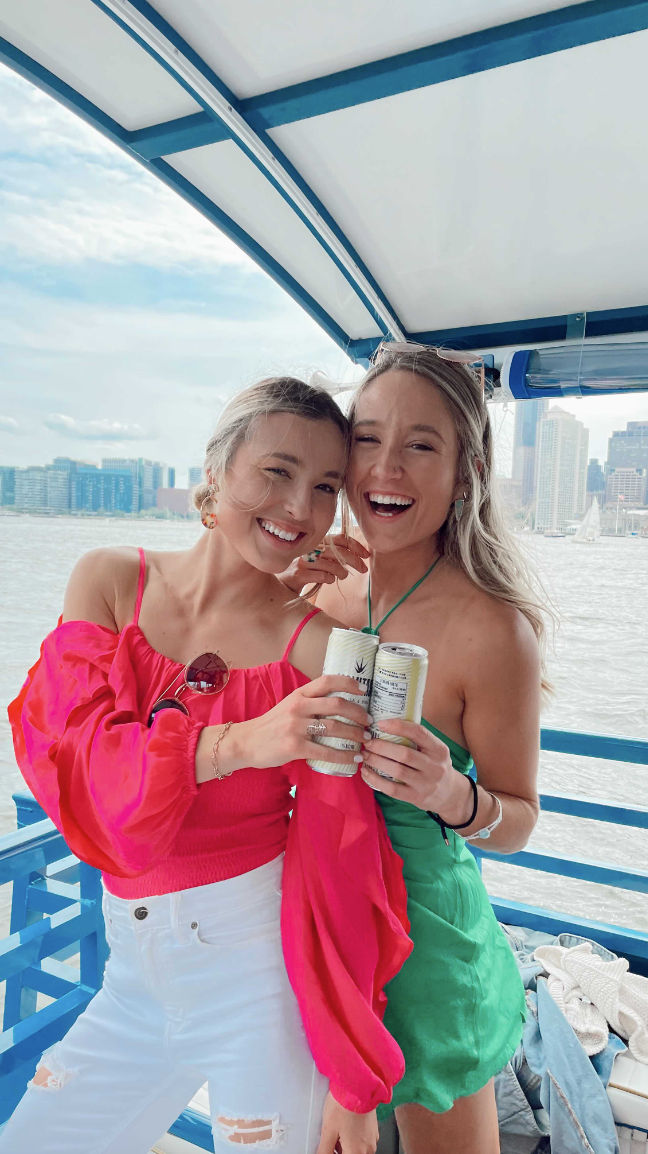 Two cheerful friends on a blue-deck boat holding canned drinks—one in a bright pink top and white ripped jeans, the other in a green sundress—smiling with an urban skyline and water in the background on a sunny waterfront boat ride.