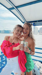 Two cheerful friends on a blue-deck boat holding canned drinks—one in a bright pink top and white ripped jeans, the other in a green sundress—smiling with an urban skyline and water in the background on a sunny waterfront boat ride.