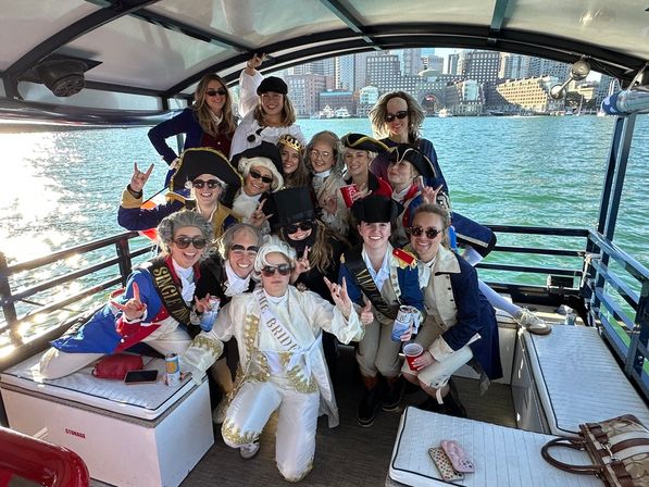 Group of friends in colonial-era costumes and sashes posing on a sunny harbor boat cruise, holding drinks with a waterfront city skyline in the background — festive bachelorette-style celebration.
