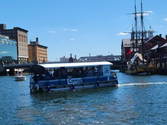 Covered sightseeing pedal boat on Boston Harbor with a yellow historic tall ship docked beside red-brick warehouses and a bridge on a sunny blue-sky day.