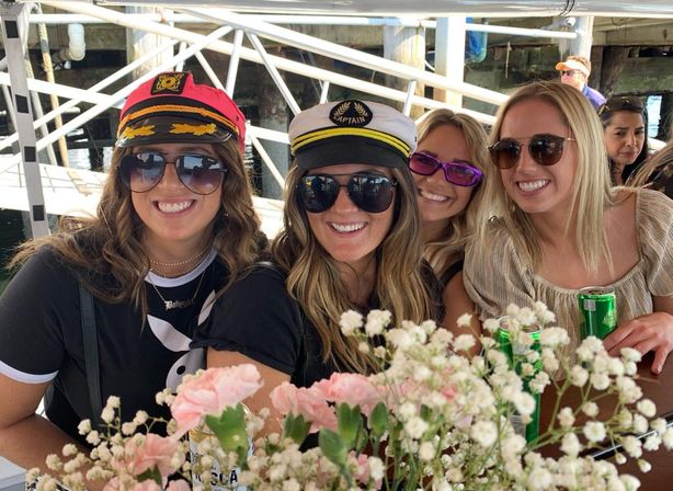 Four smiling women in captain hats and sunglasses on a docked boat at a marina, pink flowers in the foreground