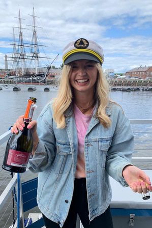Smiling woman in a captain's hat and denim jacket popping a bottle of Prosecco on a waterfront dock with a historic tall ship and brick harbor buildings in the background